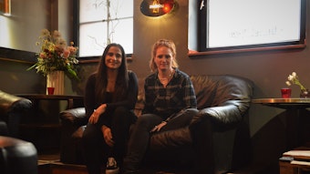 Walentini Malliou und Jana Voss sitzen auf einer Couch in der Bar ZwoEinz, über ihnen ist das Logo der Bar zu sehen.