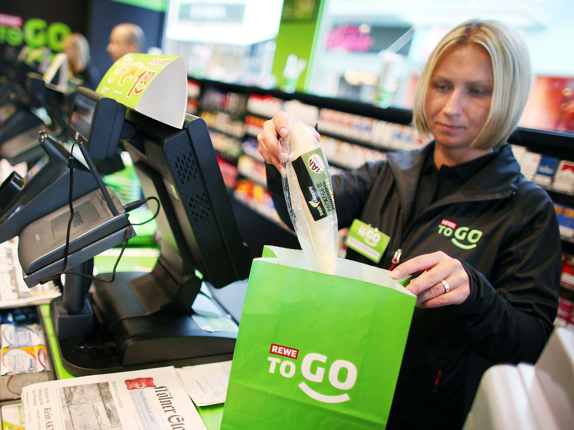 Das Symbolfoto aus dem Jahr 2011 zeigt eine blonde Kassiererin einer Rewe-To-Go-Filiale. Die Frau steht hinter der Kasse und packt Produkte in eine grüne Papiertüte.