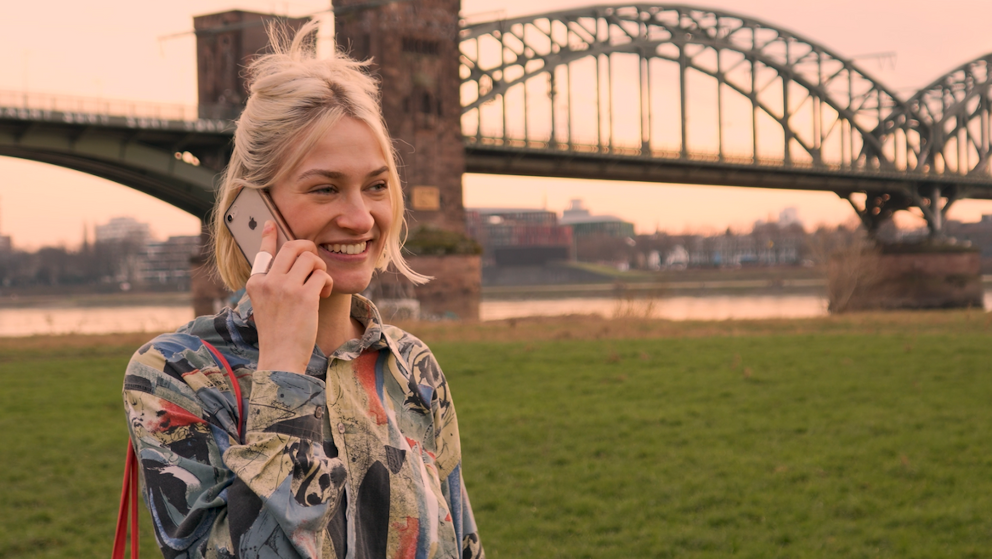 Eine junge Frau steht vor einer Kölner Brücke und telefoniert.