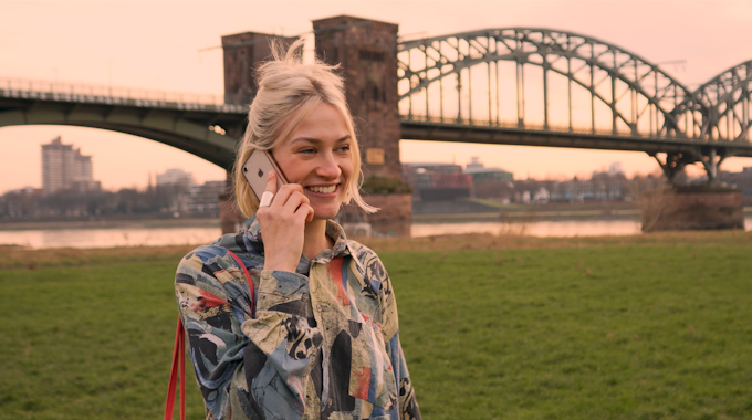 Eine junge Frau steht vor einer Kölner Brücke und telefoniert.&nbsp;