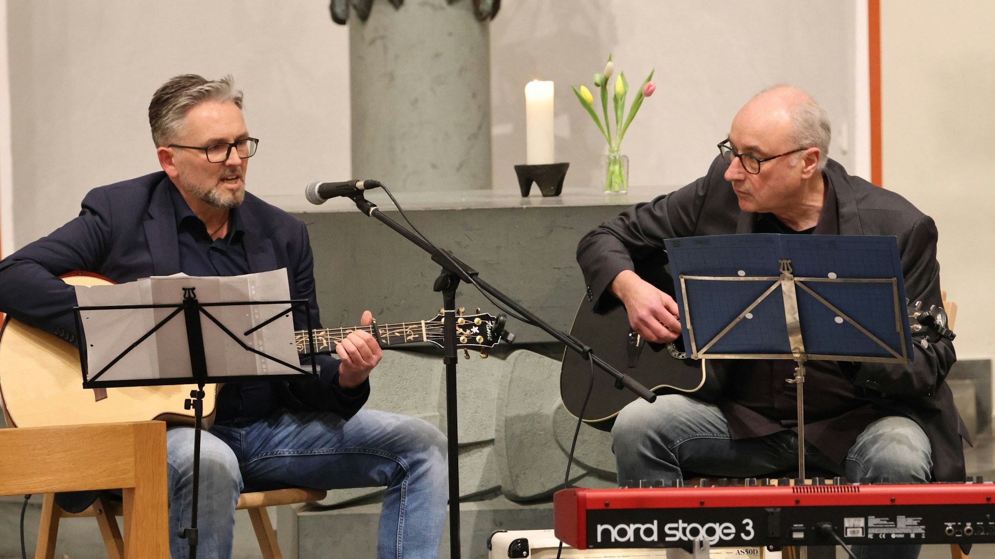 Zwei Männer sitzen in einer Kirche und spielen Gitarre. Vor ihnen stehen Notenständer.
