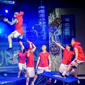 Das Bild zeigt sechs junge Männer, die akrobatisch von einem Trampolin an einen Basketballkorb springen.