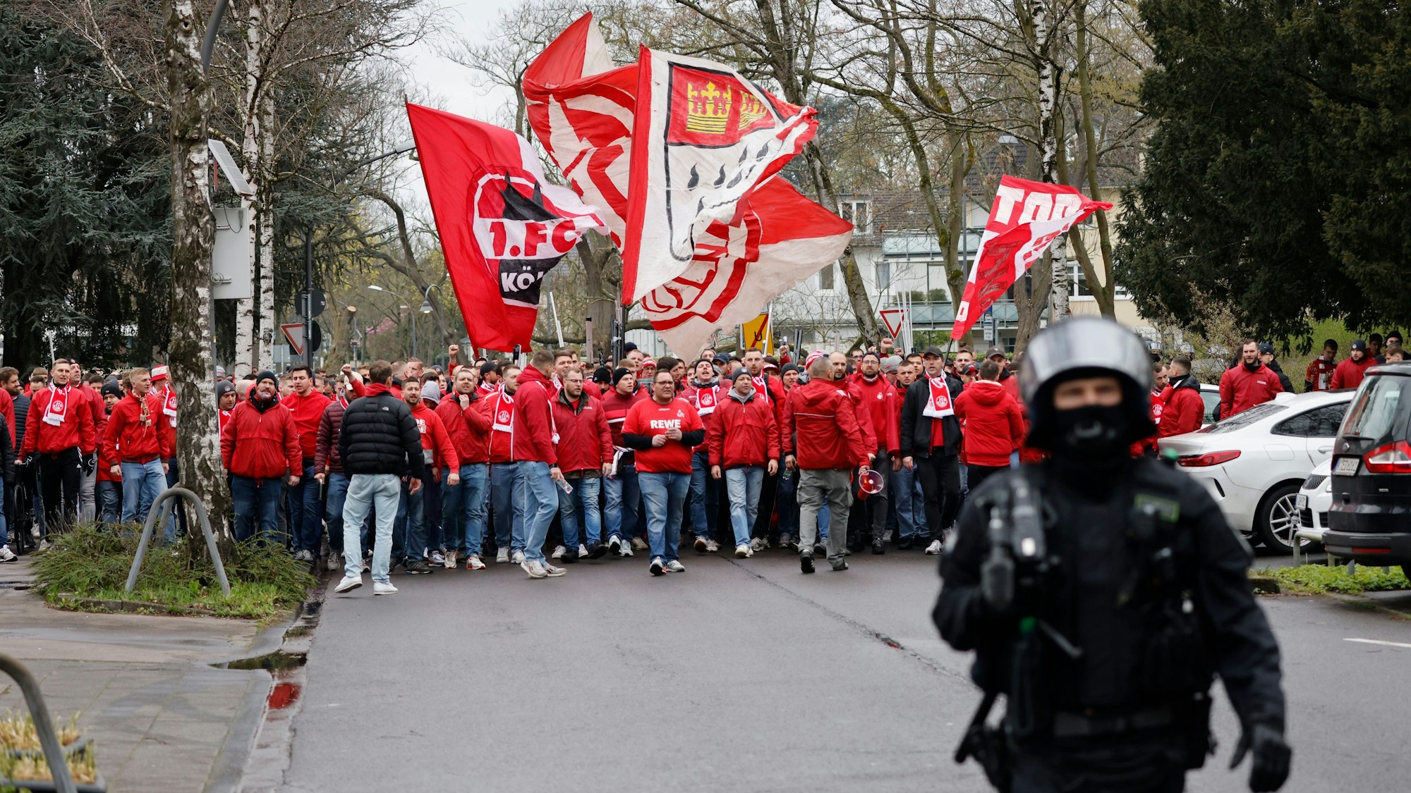 Eine große Gruppe FC-Fans mit roten Oberteilen schwenkt Fahnen und marschiert als Gruppe in Richtung des Rhein-Energie-Stadions.