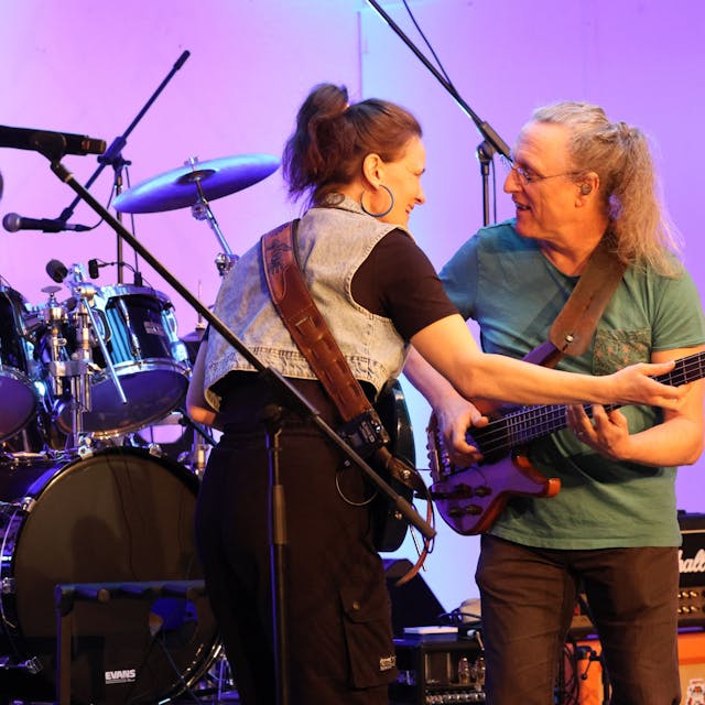 Zwei Musiker, eine Frau und ein Mann, mit Gitarre, stehen zueinander gewandt auf einer Bühne. Im Hintergrund ist ein Schlagzeuger zu sehen.