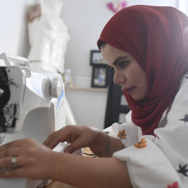Nahida Mohammadi sitzt an ihrer Nähmaschine und näht. Sie trägt eine weiße Bluse mit buntem Muster und ein dunkelrotes Kopftuch.