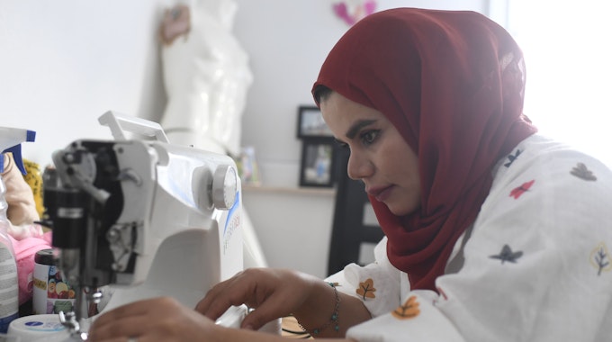 Nahida Mohammadi sitzt an ihrer Nähmaschine und näht. Sie trägt eine weiße Bluse mit buntem Muster und ein dunkelrotes Kopftuch.