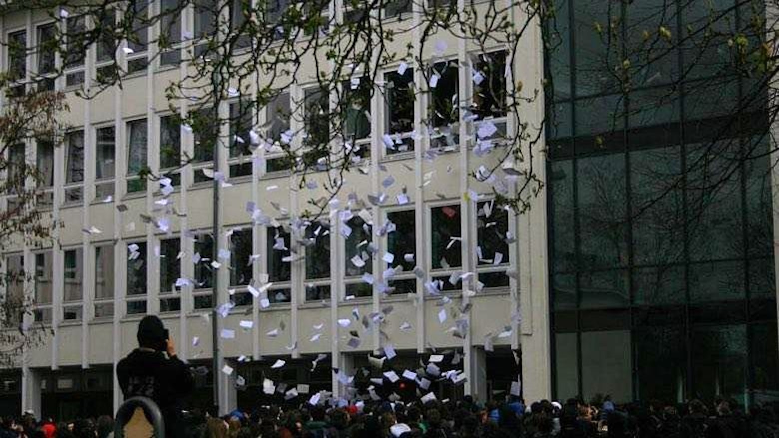 Tausende bedruckte Blätter fliegen aus Fenstern eines Schulgebäudes. Vor dem Gebäude steht eine große Gruppe Schüler.
