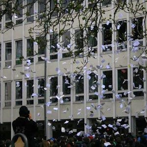 Tausende bedruckte Blätter fliegen aus Fenstern eines Schulgebäudes. Vor dem Gebäude steht eine große Gruppe Schüler.