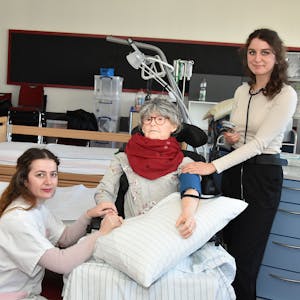 Zwei junge Frauen stehen in einem Krankenzimmer. In ihrer Mitte ist eine Puppe mit dem Aussehen einer älteren Frau, an der sie den Patientenkontakt üben.&nbsp;
