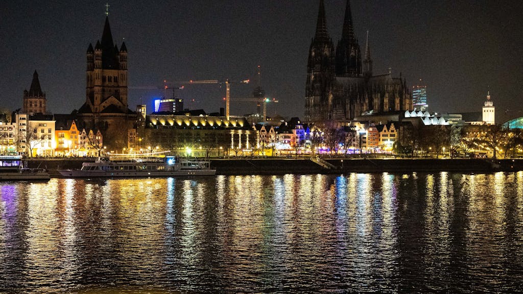 Die beleuchtete Rheinpromenade in Köln in der Dunkelheit. Im Hintergrund ist der Kölner Dom zu sehen, der nicht beleuchtet ist.