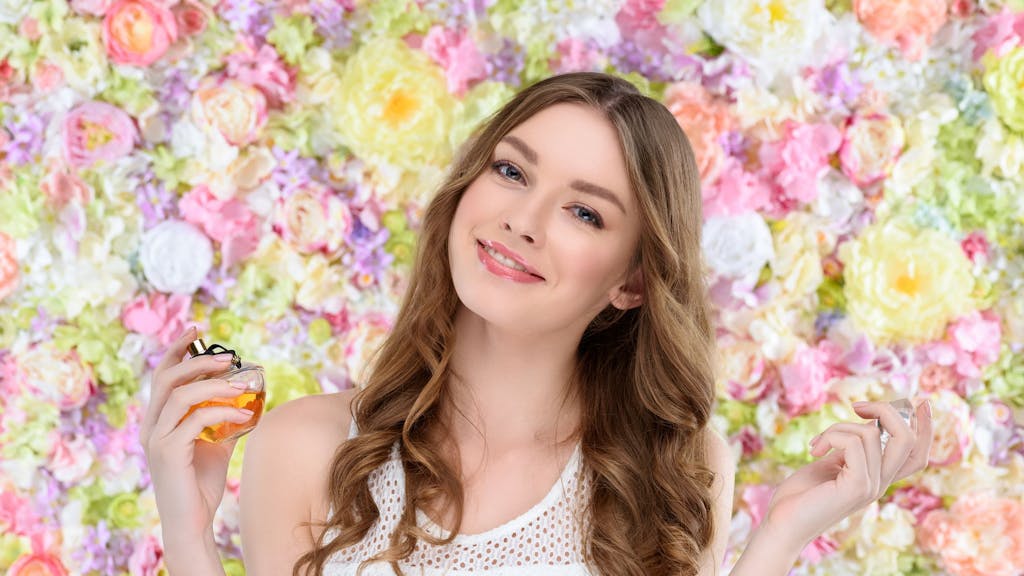 Junge Frau sprüht sich vor einem floralen Frühlings-Hintergrund Parfüm aufs Handgelenk.