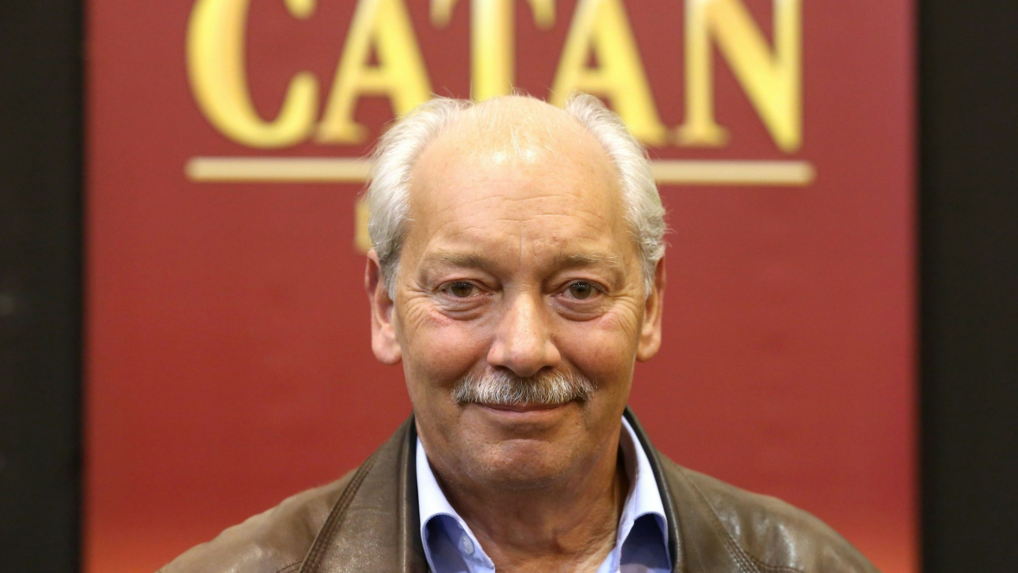Klaus Teuber, der Erfinder des weltberühmten Strategiespiels „Die Siedler von Catan“, steht vor einer Wand mit der Aufschrift „Catan“.