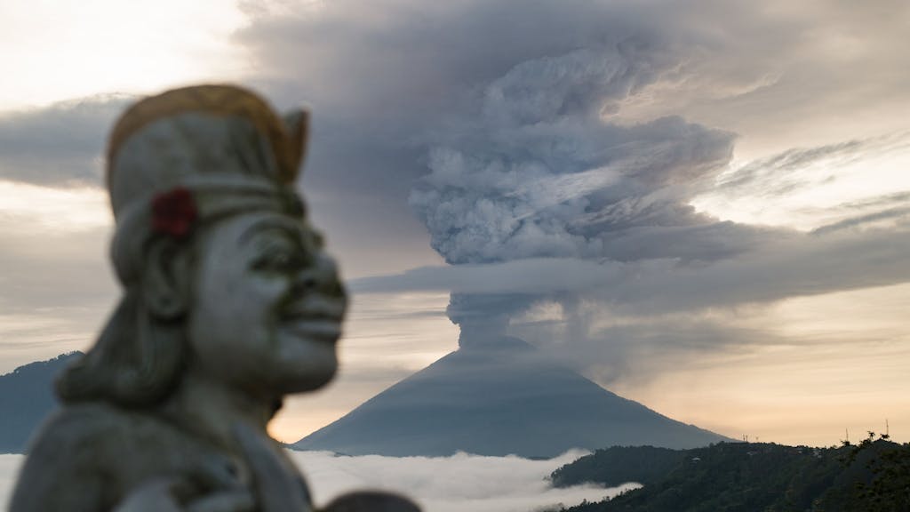 Der heilige Berg Agung auf der indonesischen Insel Bali während eines Vulkanausbruches. Rauch steigt aus dem Vulkan empor. Im Vordergrund ist eine Statue zu sehen.
