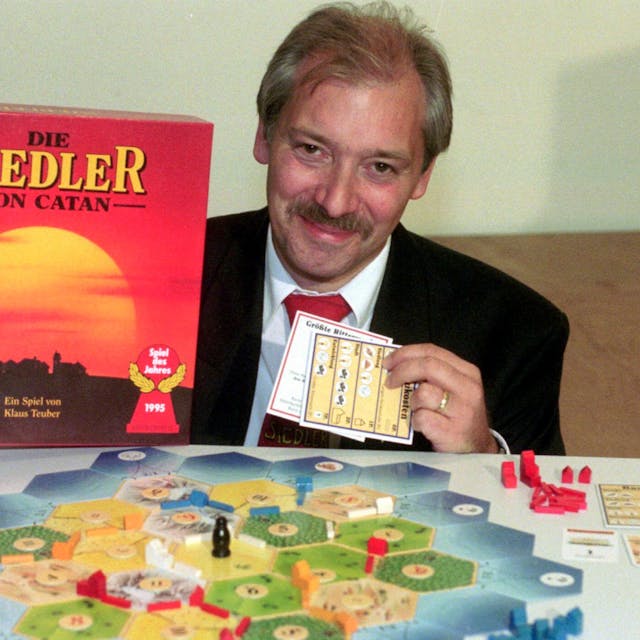 Klaus Teuber sitzt an einem Tisch, in der Hand hält der die Spielschachtel von „Catan“, vor ihm ist das Spiel aufgebaut.