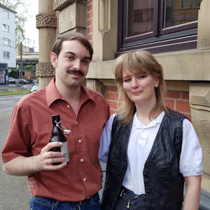 Die beiden sind gekleidet wie in den 60er-Jahren. Luca trägt einen Schnurbart und hält ein Bier in der Hand. Sabine trägt ein weißes Top mit einer schwarzen Weste.