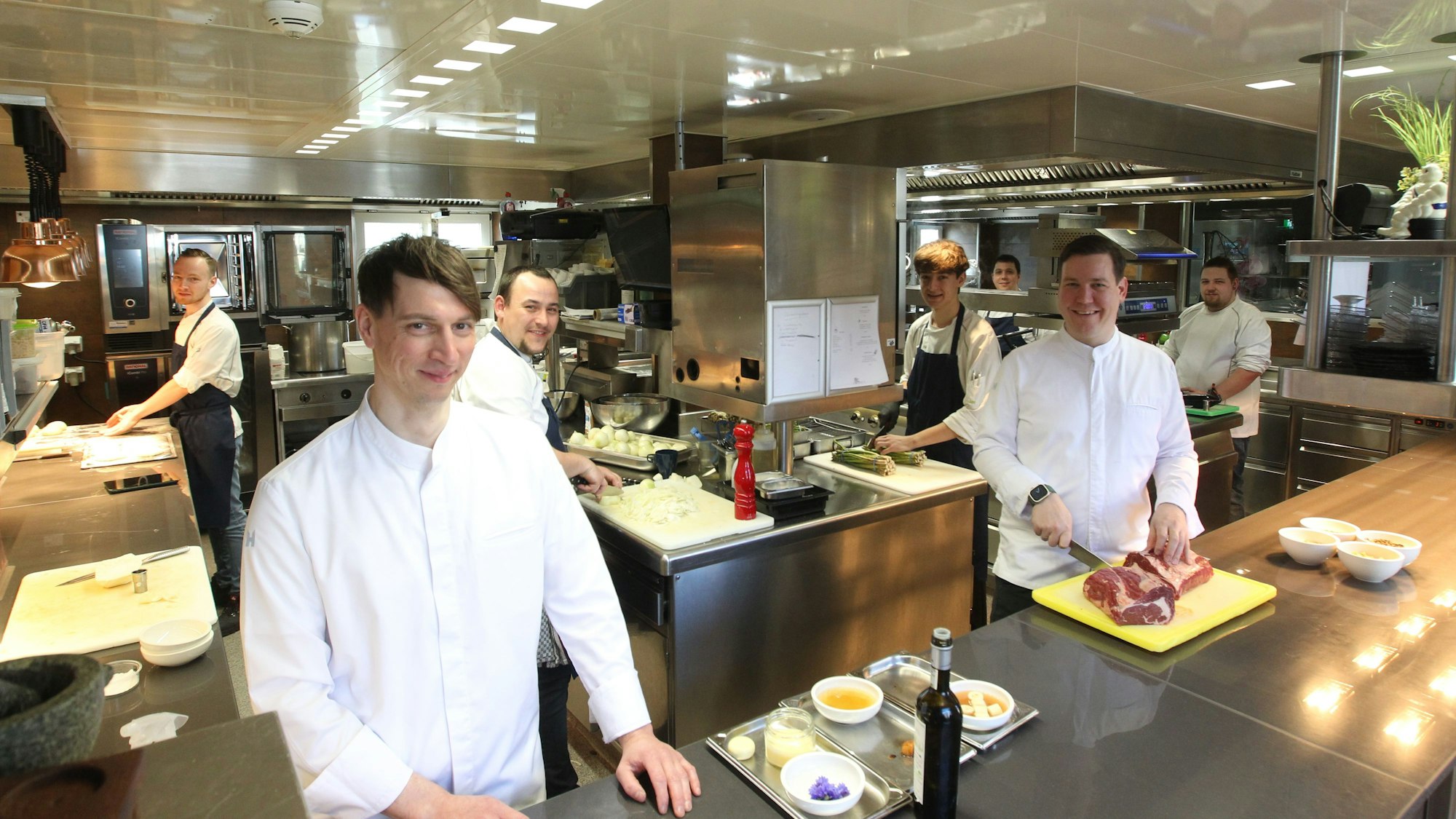 Thomas und sein Team lächeln in die Kamera und stehen an verschiedenen Stationen in der Küche.