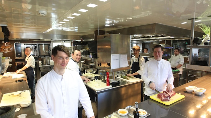 Thomas und sein Team lächeln in die Kamera und stehen an verschiedenen Stationen in der Küche.&nbsp;
