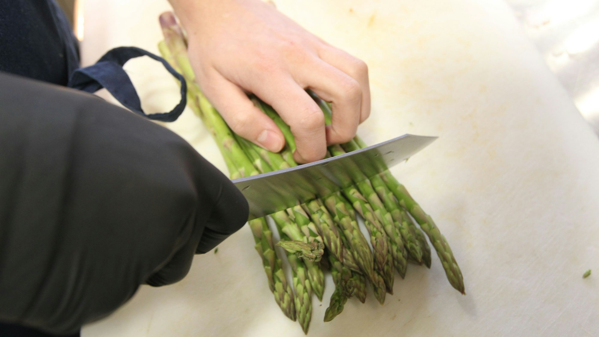 Grüner Spargel wird geschnitten. Die Hand mit dem Messer trägt einen schwarzen Handschuh.