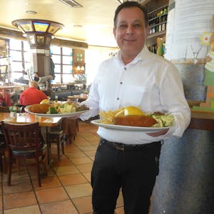 Iakovos Sidiropoulos, Geschäftsführer des „Gilden Brauhaus“ in Köln-Mülheim, steht in seinem Restaurant und hat zwei Teller mit Speisen in den Händen.
