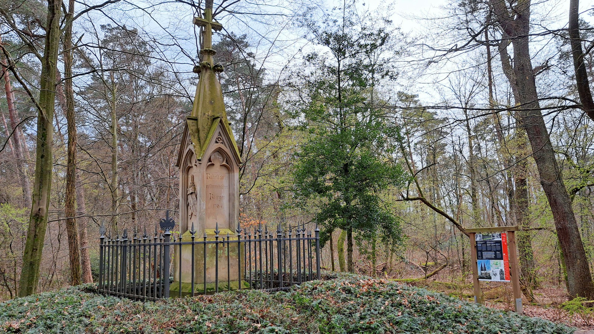 Ein gotisches Denkmal steht auf einem Hügel. Rechts im Hintergrund ist eine bunter Informationstafel zu sehen.