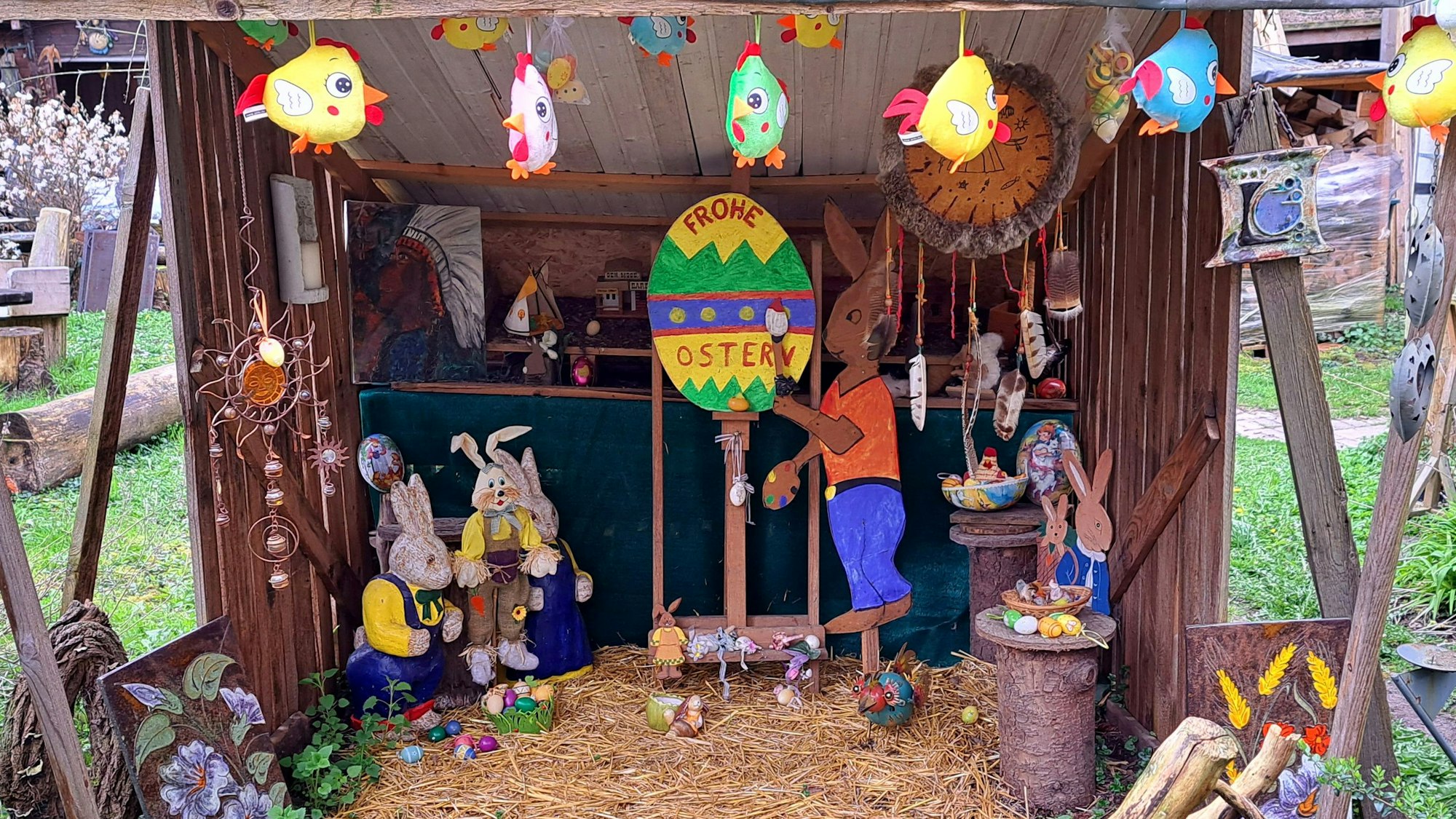 Hasenfiguren stehen in einem Unterstand, auf einem großen bunten Osterei steht „Frohe Ostern“.