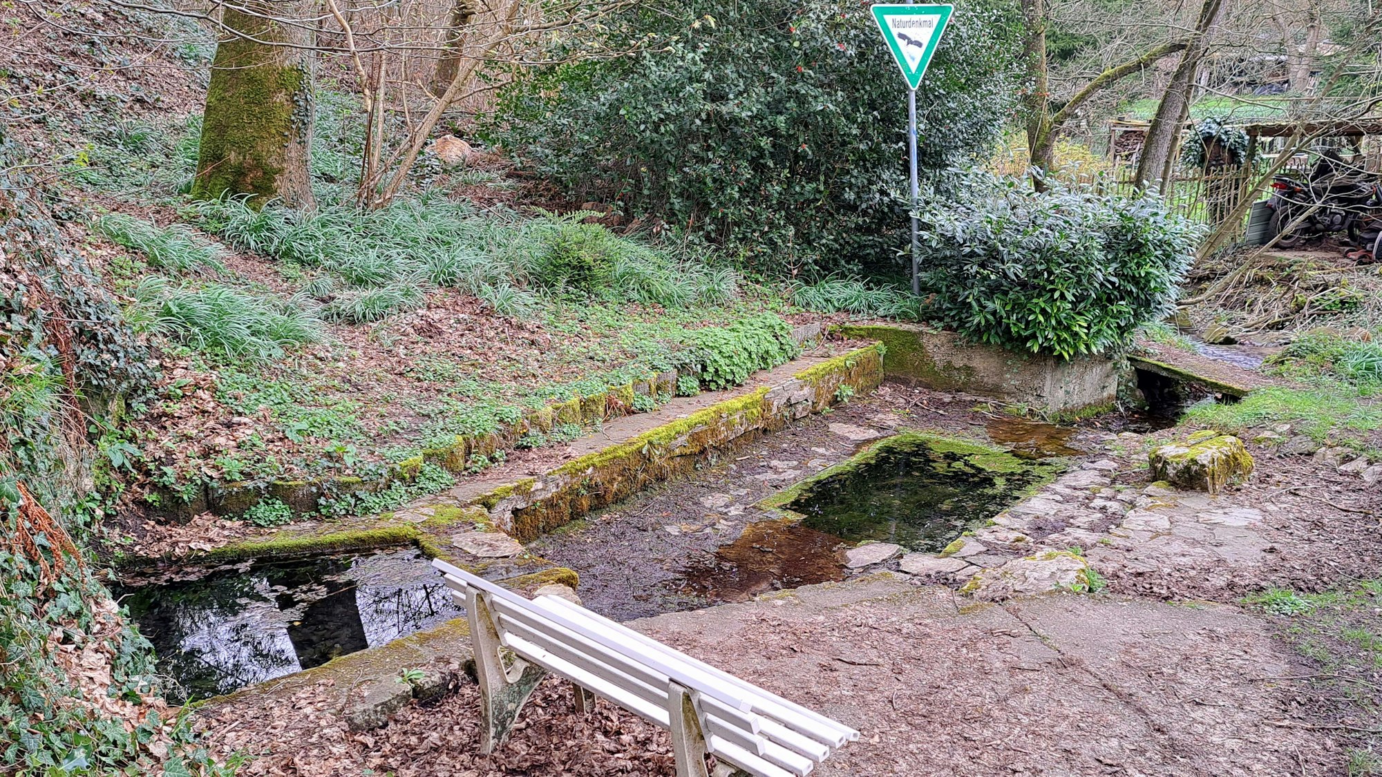 Neben einer mit Mauern eingefassten Wasserstelle steht eine Bank und ein Naturdenkmal-Schild.