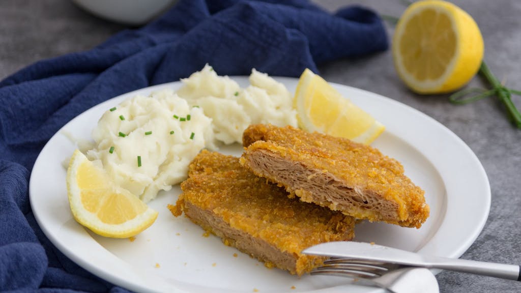 Auf dem Foto sieht man ein veganes Schnitzel auf einem Teller, zusammen mit Kartoffelpüree und einer Zitrone.