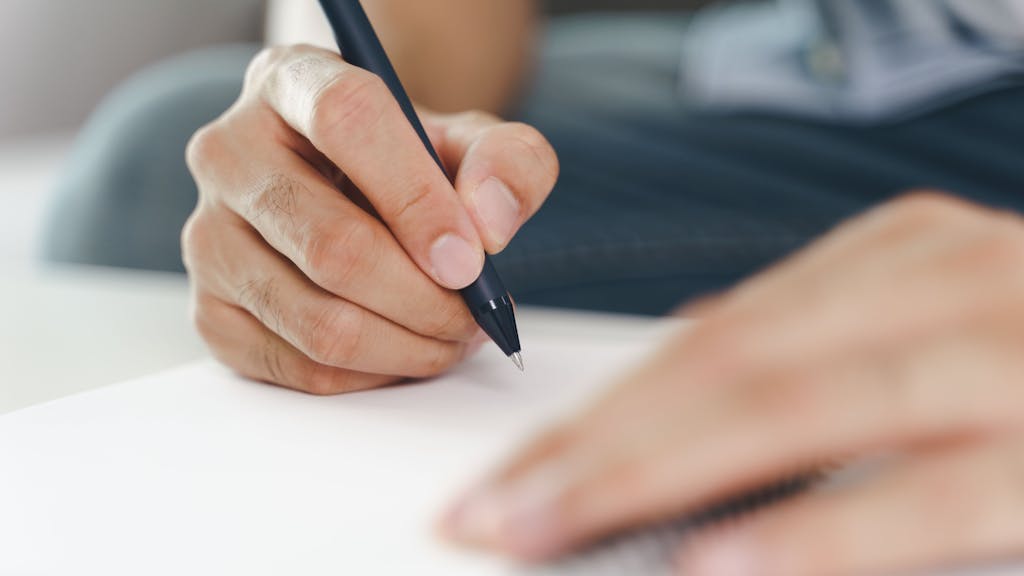 Close up of young man in casual cloth hands writing down on the notepad, notebook using ballpoint pen on the table.