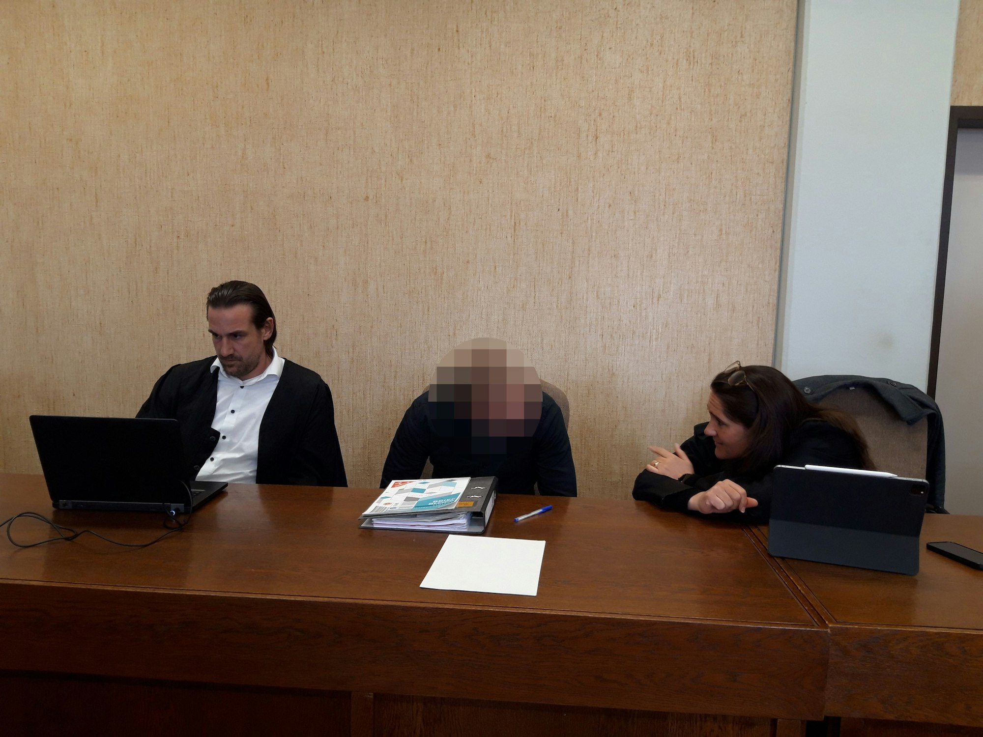 In einem Saal im Kölner Landgericht sitzt eine Person, deren Gesicht unkenntlich ist, zwischen einem Mann, der an einem Laptop arbeitet, und einer Frau, die mit ihm ins Gespräch vertieft ist.