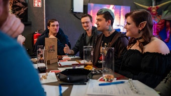 Eine Gruppe junger Menschen sitzt an einem Tisch und spielt ein Rollenspiel, vor ihnen liegen Würfel. Eine von ihnen trägt Elfenohren.