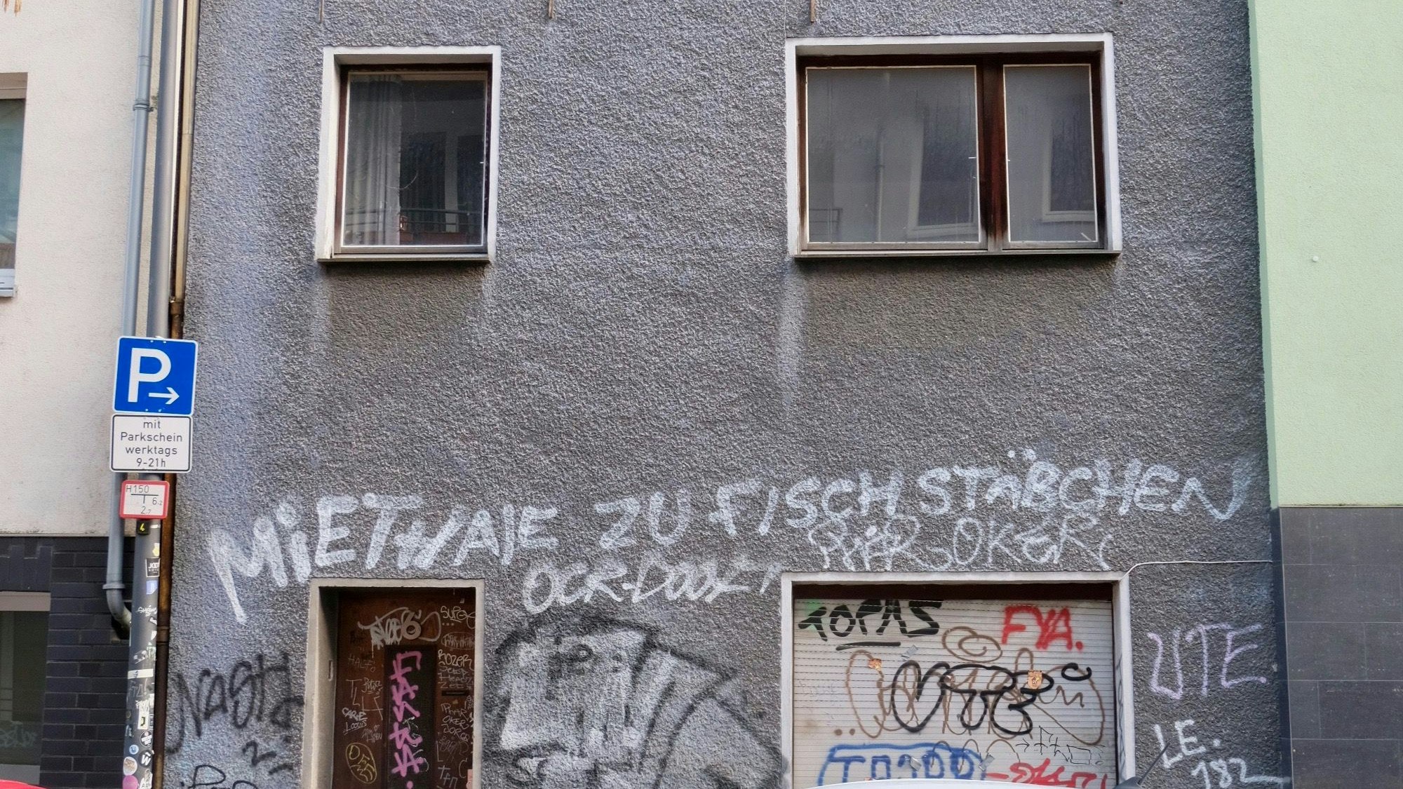 Ein runtergekommenes, unbewohntes Haus in der Kölner Glasstraße 6, mit grauer Fassade, auf die der Spruch „Miethaie zu Fischstäbchen“ gesprüht ist, steht seit mindestens 15 Jahren leer.