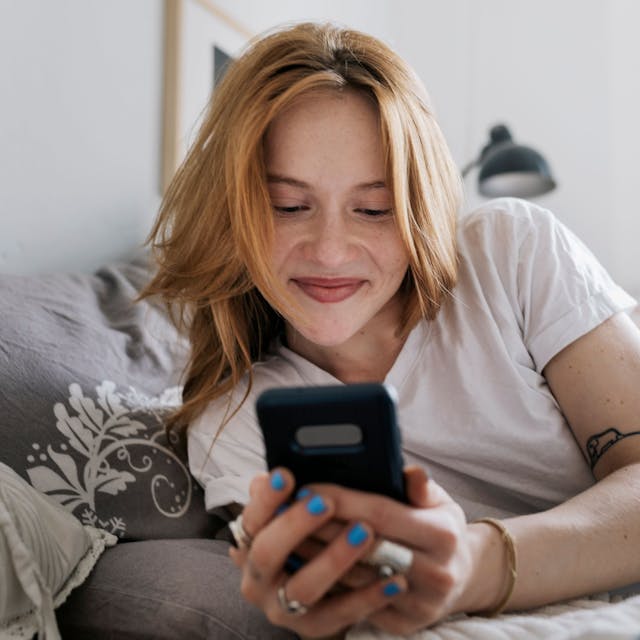 A woman smiling while lying on her bed at home and messaging on an online dating app using her smartphone.