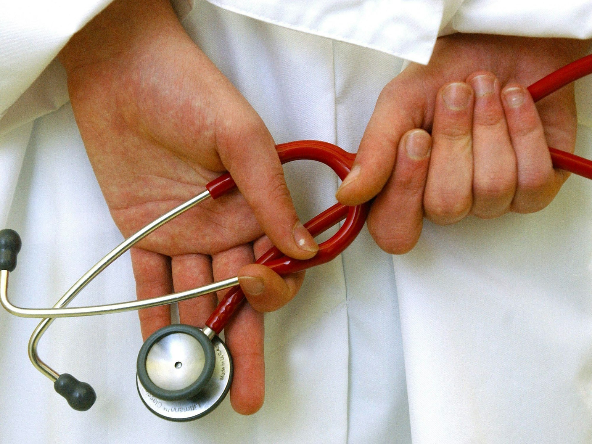 Hier zu sehen, die Hände eines Arztes in weißem Kittel. Er hält ein rotes Stethoskop.