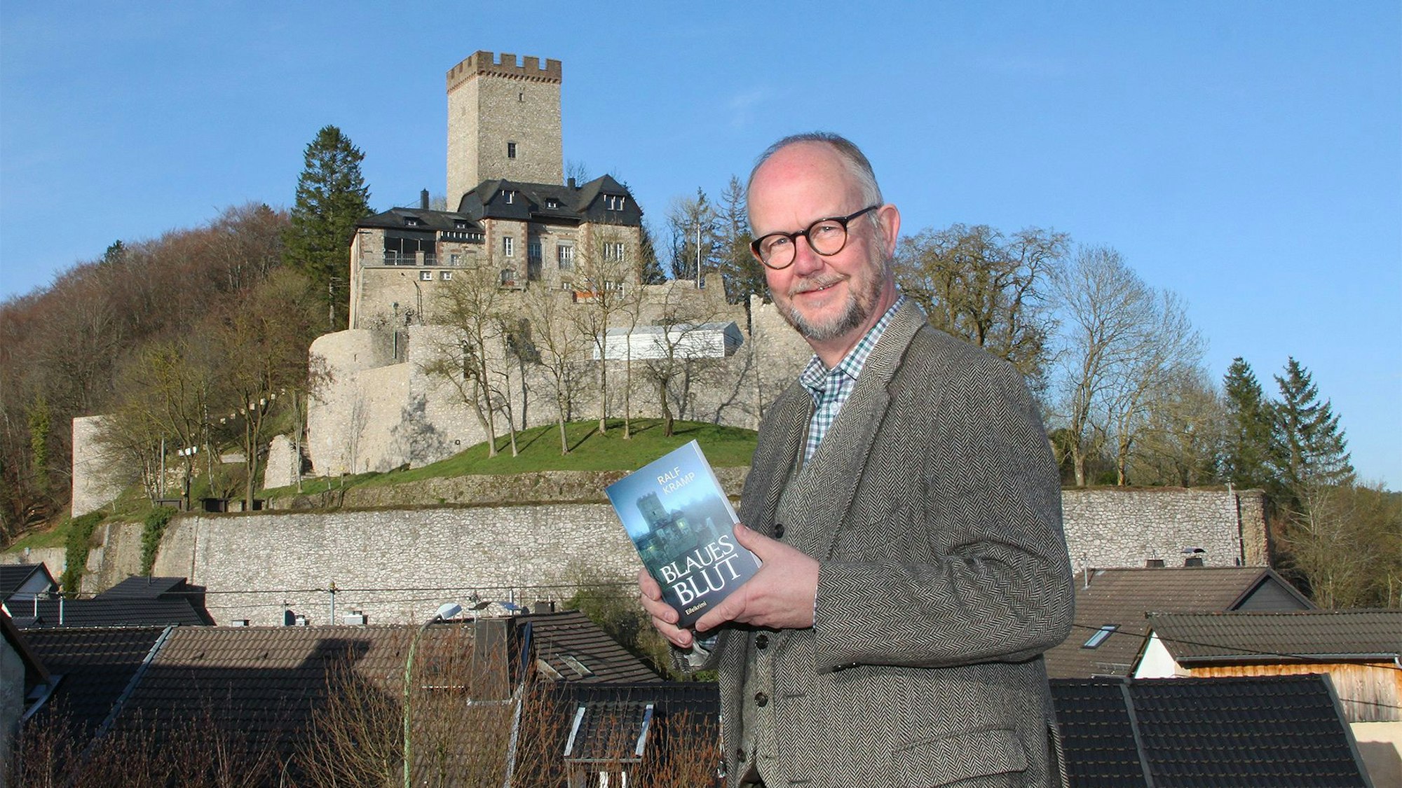 Krimiautor Ralf Kramp steht vor der Burg Kerpen, die auch auf dem Cover seines neuen Buches zu sehen ist, das er in der Hand hält.