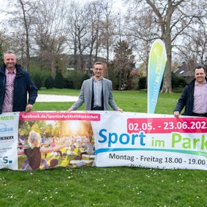 Die drei Männer posieren mit dem Banner von „Sport im Park“ und lächeln in die Kamera.&nbsp;