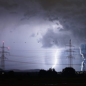 Zwei Blitze schlagen in der Nähe eines Strommastes ein, der Himmel ist stark verdunkelt. Im Hintergrund sind einige Häuser zu sehen. (Symbolbild)