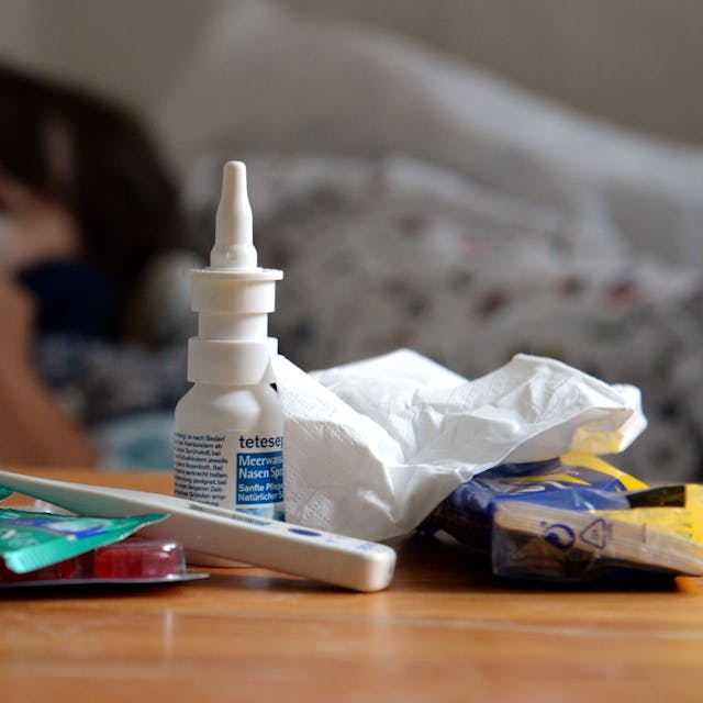 Wer Grippe-Symptome hat, der sollte auf jeden Fall das Bett hüten und nicht zur Arbeit gehen, betonen Ärztinnen und Ärzte. Unser Foto zeigt einen kranken Menschen im Bett und einen Stapel von Medikamenten.