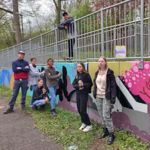 Die Graffiti-Künstler stehen mit dem ehrenamtlichen Workshop-Leiter Hannes Fries an der besprühten Mauer neben der Opladener Straße.