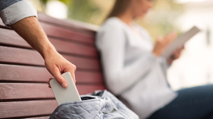 Symbolfoto: Dieb stiehlt Smartphone aus der Tasche einer Frau, die auf einer Parkbank sitzt und liest.