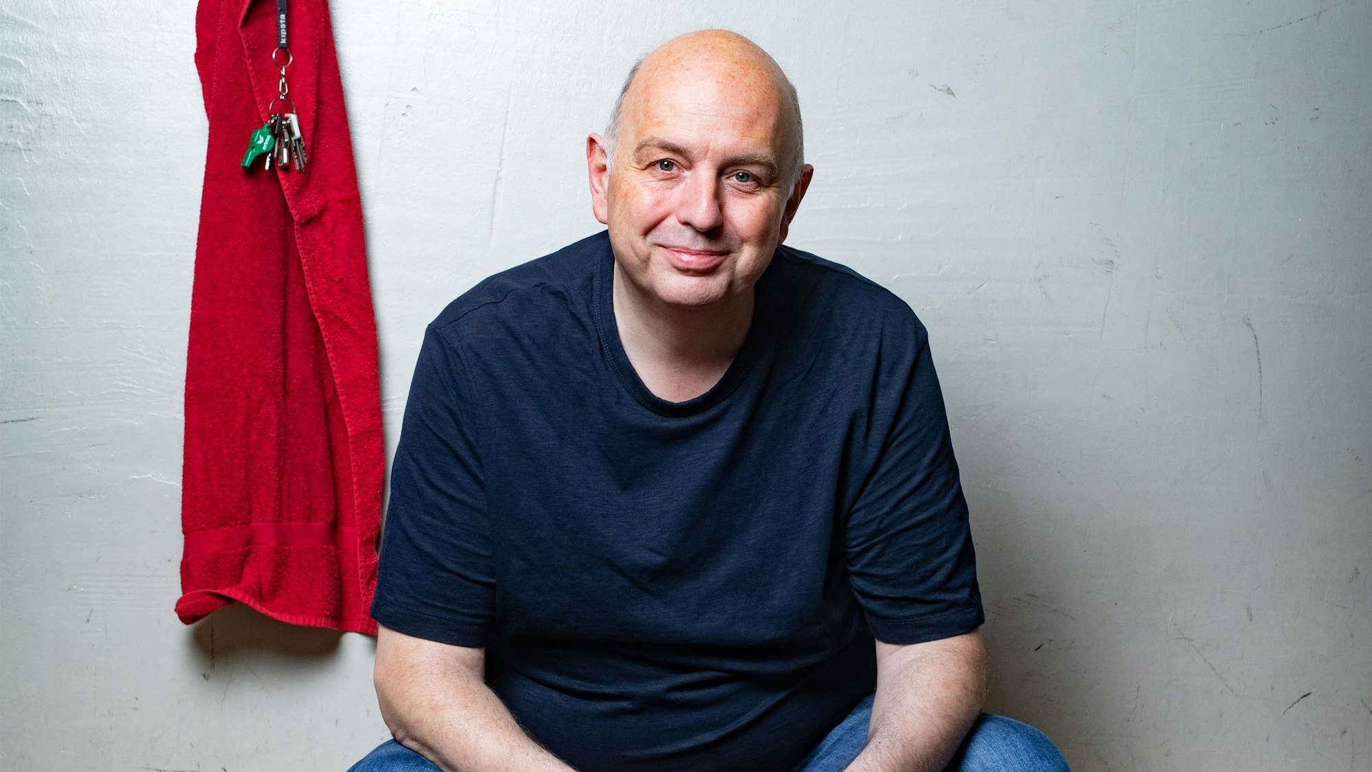 Frank Goosen sitzt auf einer Bank, in einem blauen T-Shirt vor einer weißen Wand. Links von ihm hängt ein rotes Handtuch und ein Schlüsselbund mit Trillerpfeife.