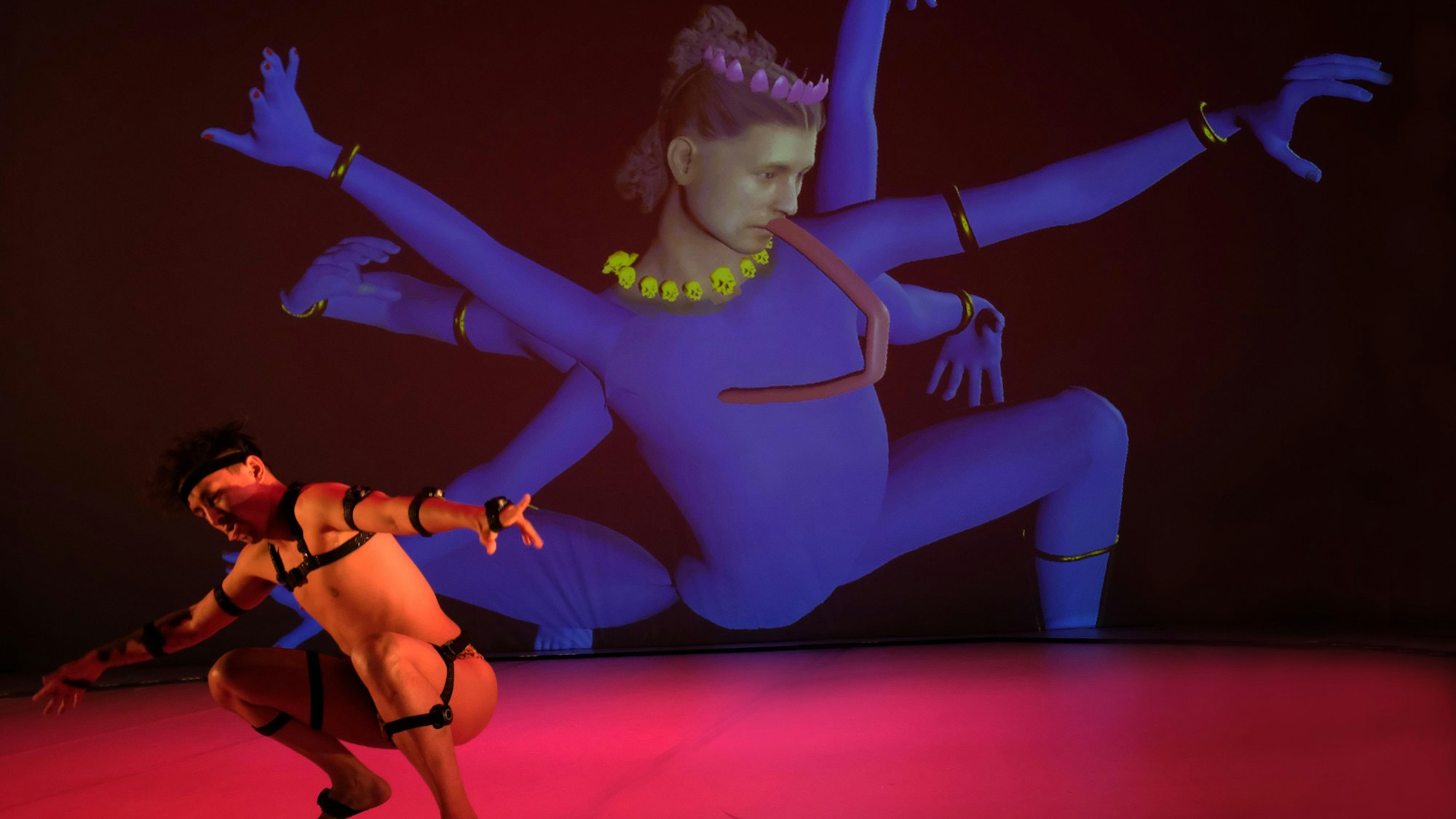 Ein Tänzer aus Singapur in Lederriemen tanzt vor einer Videoinstallation, die eine singapurische Gottheit persifliert.