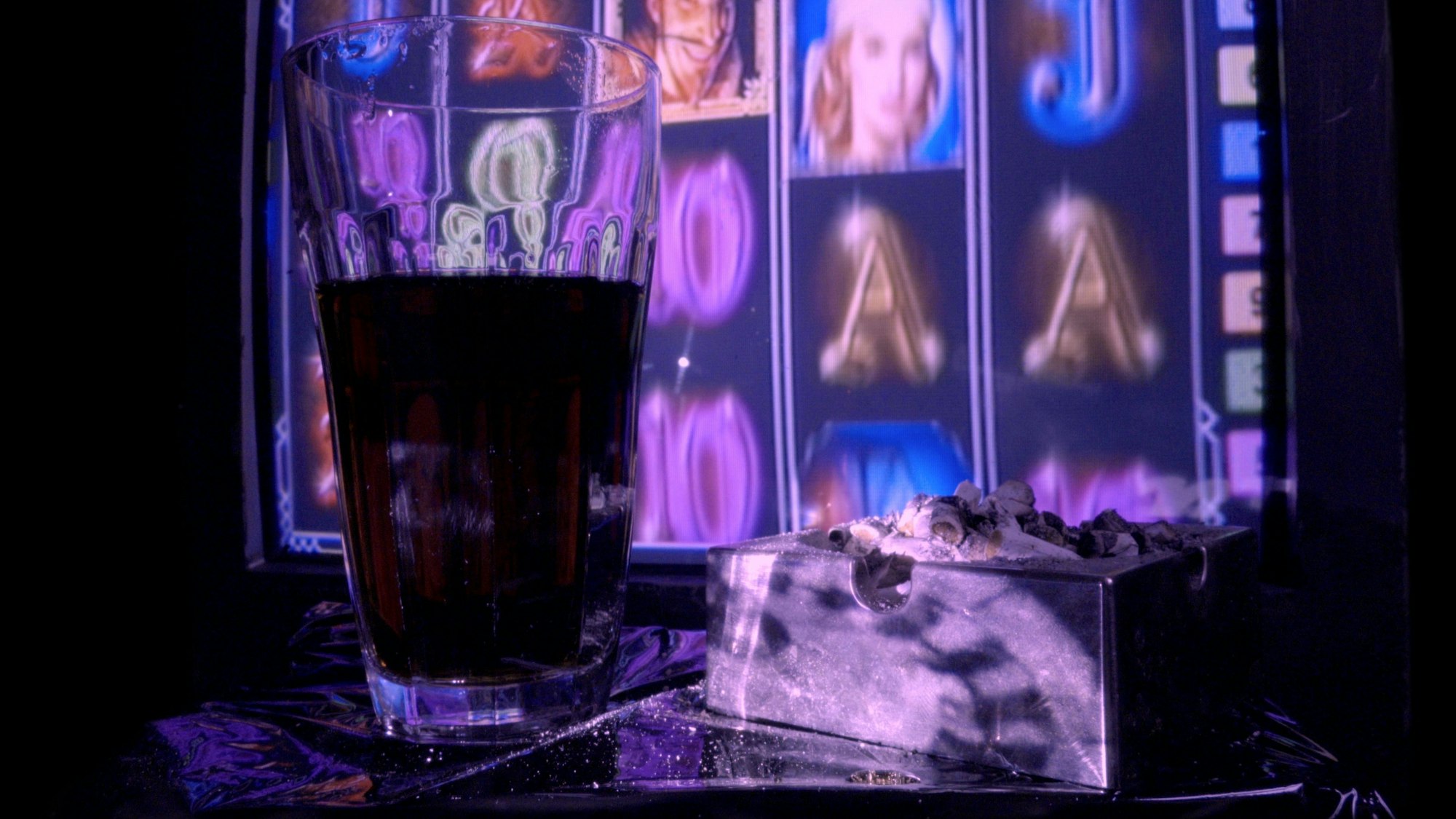 Vor einer Videoinstallation, die einen Glückspielautomaten zeigt, stehen ein Glas mit Cola und ein voller Aschenbecher.