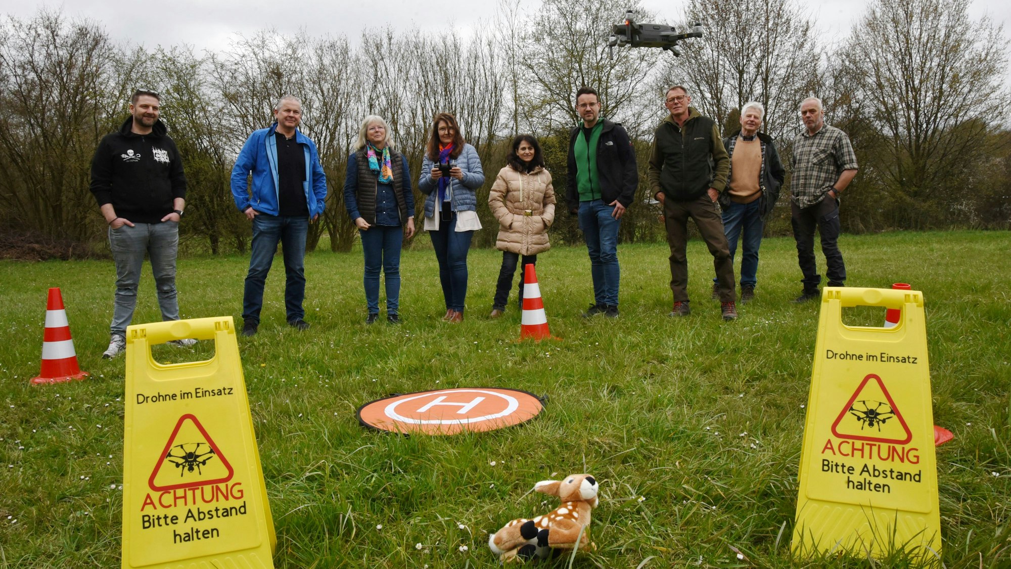Mehrere Leute stehen auf einer Wiese in einer Reihe. Einer fliegt mit einer Drohne über ein im Gras liegendes Stoff-Rehkitz. Daneben stehen zwei gelbe Warnschilder mit der Aufschrift: „Drohne im Einsatz. Achtung, bitte Abstand halten“.