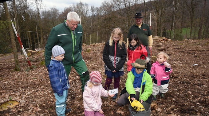 Mehrere Kinder pflanzen unter Anleitung eines älteren Mannes in grünem Overall kleine Setzlinge.