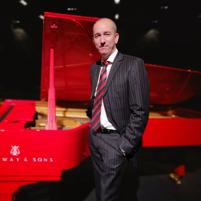 Der amerikanische Pianist Chris Hopkins spielt am 1. Mai in der Ülepooz. Er steht im Anzug vor einem roten Flügel.