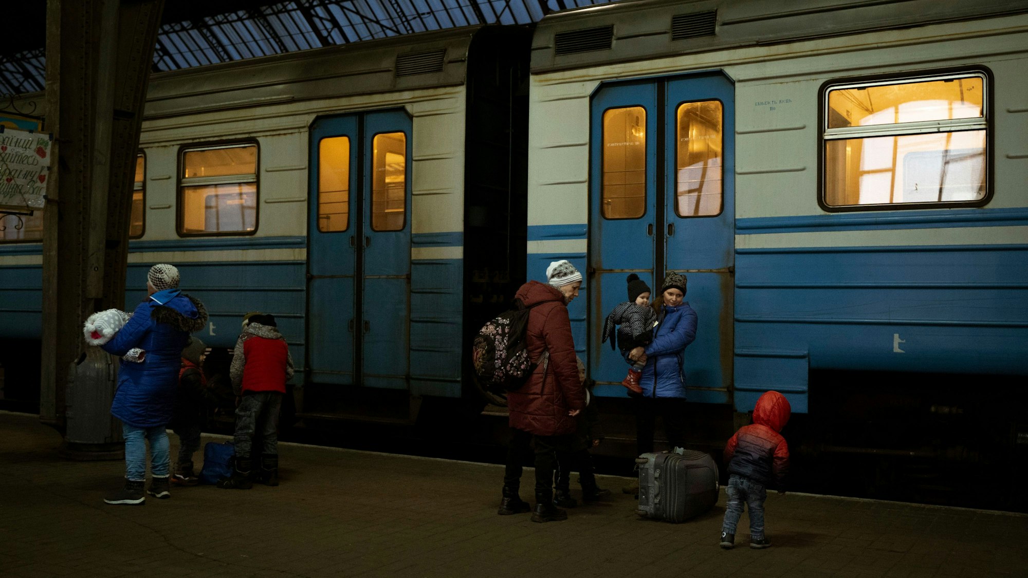 Zu sehen sind Frauen mit ihren Kindern. Am Gleis steht ein blau-weiß gestreifter Zug.