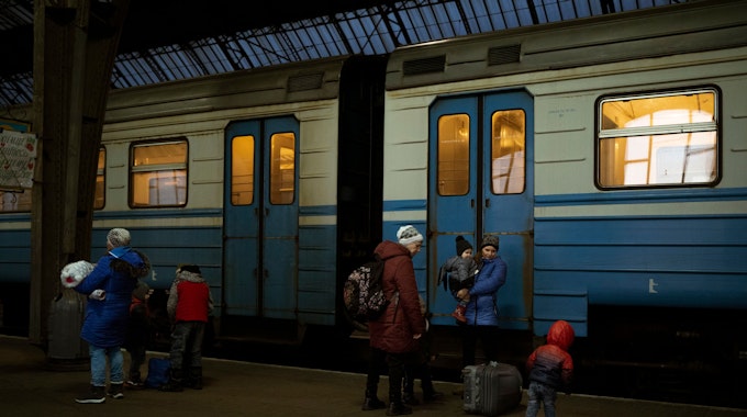 Zu sehen sind Frauen mit ihren Kindern. Am Gleis steht ein blau-weiß gestreifter Zug.&nbsp;