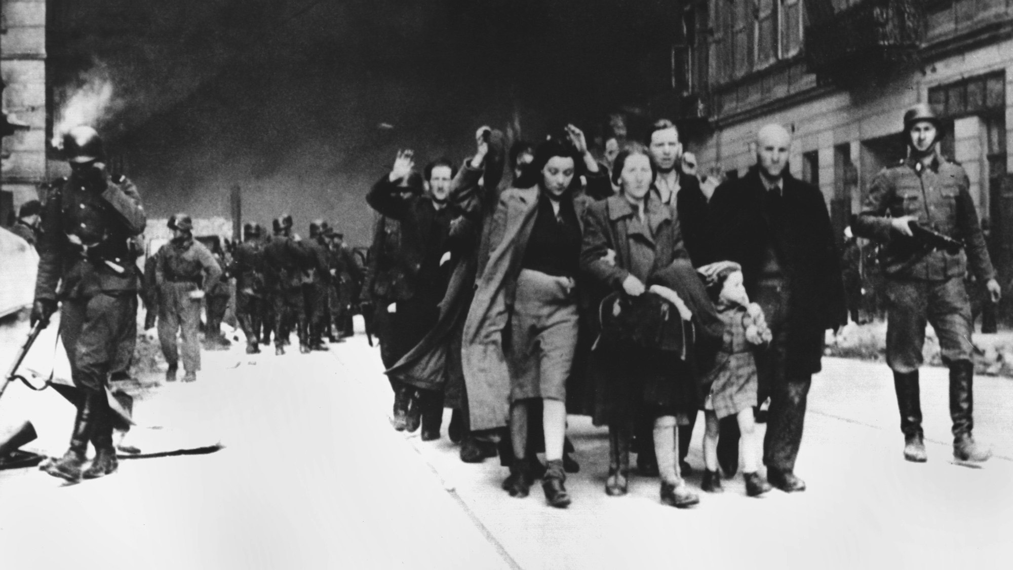Mit erhobenen Händen werden im Mai 1943 jüdische Frauen, Männer und Kinder von deutschen SS-Soldaten aus dem brennenden Ghetto der polnischen Hauptstadt Warschau getrieben. Die Aufnahme entstand in der letzten Phase des Ersten Warschauer Aufstandes, der vom 19. April 1943 bis zu seiner blutigen Niederschlagung am 16. Mai 1943 dauerte.