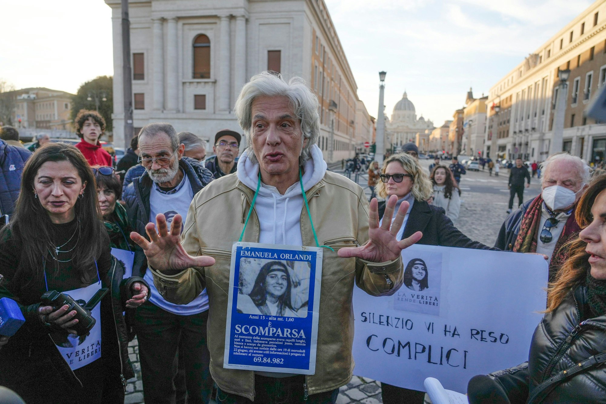 Emanuelas Bruder Pietro Orlandi sucht im Vatikan nach seiner verschwundenen Schwester. Um den Hals hängt das Fahndungsplakat mit Emanuela Orlandis Foto. Um ihn herum stehen weitere Demonstrierende, die Aufklärung fordern. Im Hintergrund sieht man den Petersdom.