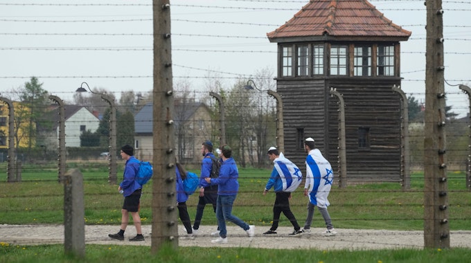 Das Symbolfoto aus dem Jahr 2022 zeigt einen Teil des Konzentrationslagers Auschwitz. Im Vordergrund befindet sich ein Stacheldrahtzaun, dahinter laufen einige Menschen in blauen Jacken und mit der israelischen Flagge.
